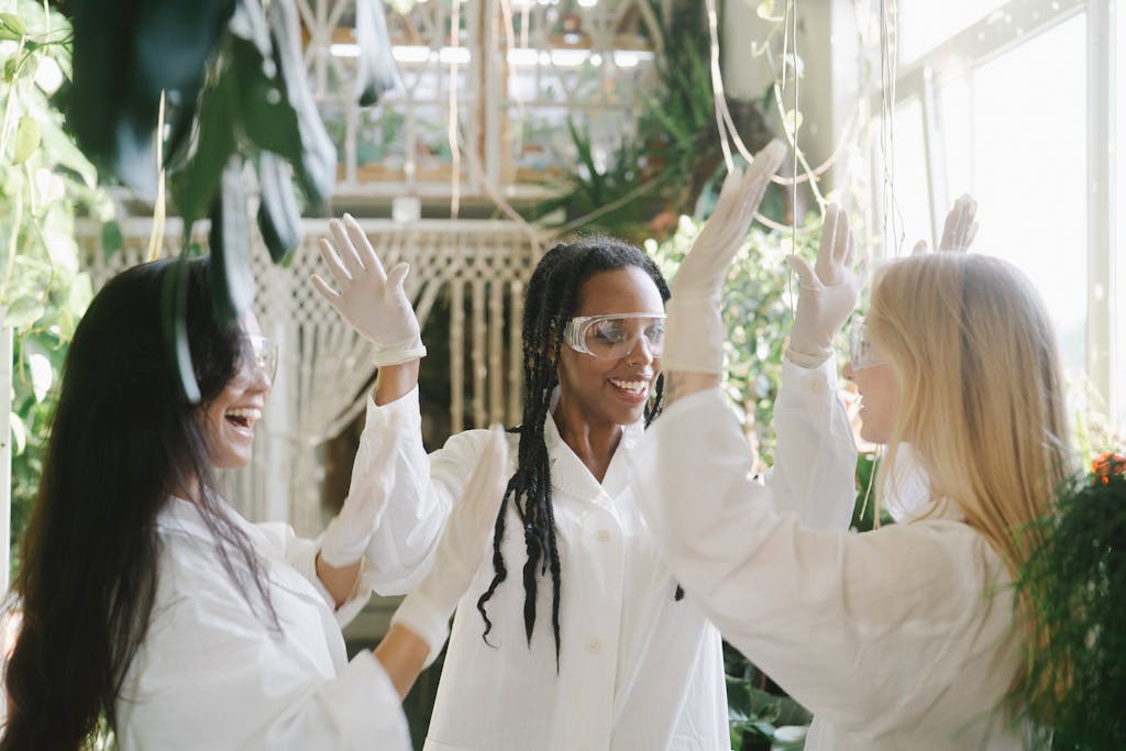 Three female scientists in lab gowns and goggles high-five happily in a plant-filled indoor laboratory.